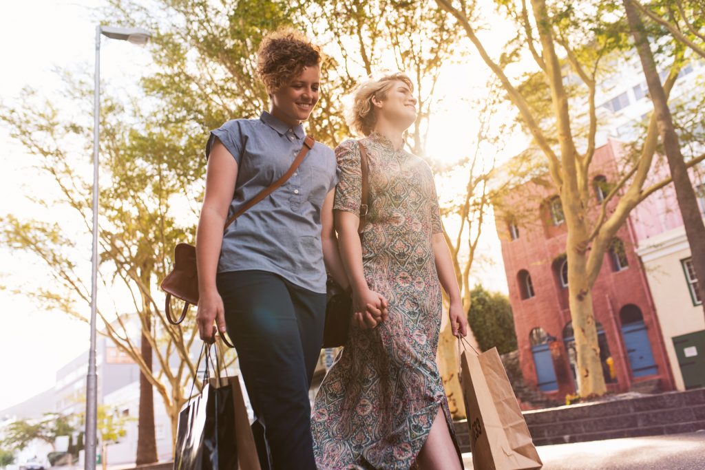 Smiling young lesbian couple walking hand in hand together down a city street on a sunny day carrying shopping bags